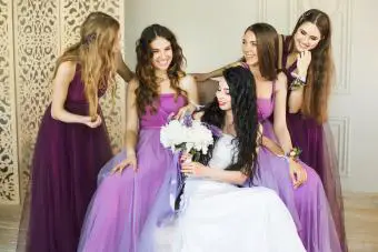 Novia con ramo de novia y sonriendo mientras habla con sus damas de honor en vestidos morados 