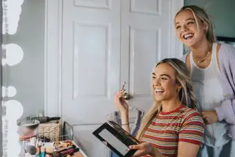 Dos mujeres jóvenes se ríen frente a un espejo iluminado y se hacen un cambio de imagen.