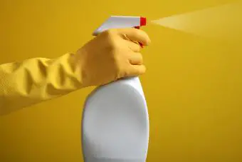 La mano en guante de goma amarillo sostiene una botella de spray de plástico con detergente de limpieza sobre fondo amarillo.