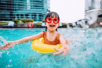 Niña juguetona con gafas de sol sonriendo alegremente, salpicando y jugando con agua alegremente en la piscina en las vacaciones de verano